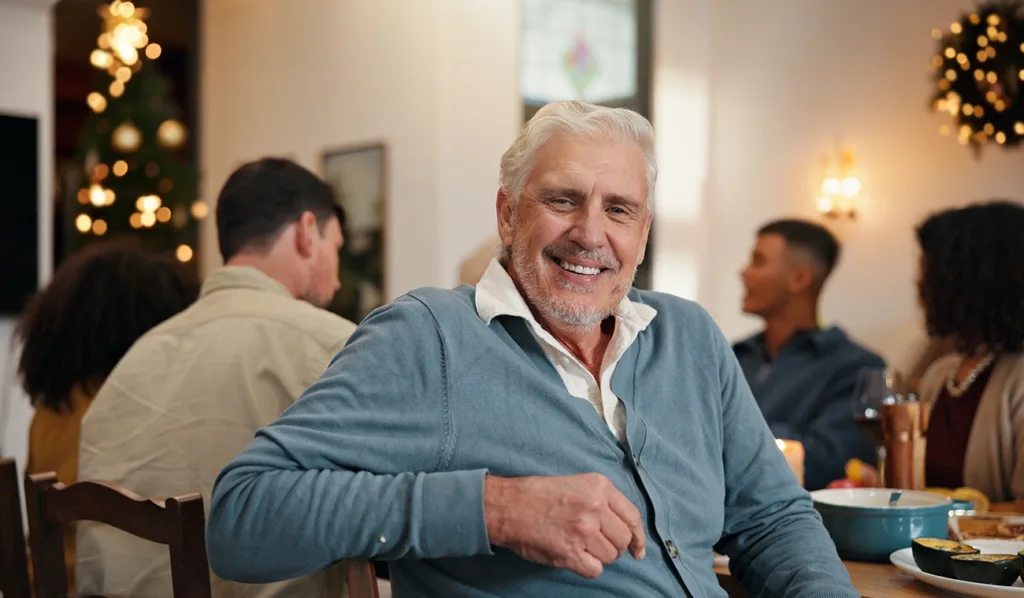 An older man with gray hair, smiling and sitting at a dinner table with his family enjoying a holiday celebration with festive lights and decorations out of focus in the background